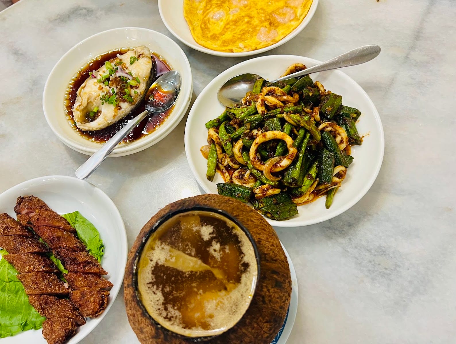 Steamed fish in soy sauce, stir-fried squid with okra and long beans, a prawn omelette, fried meat rolls and a coconut-shell soup on a marble table