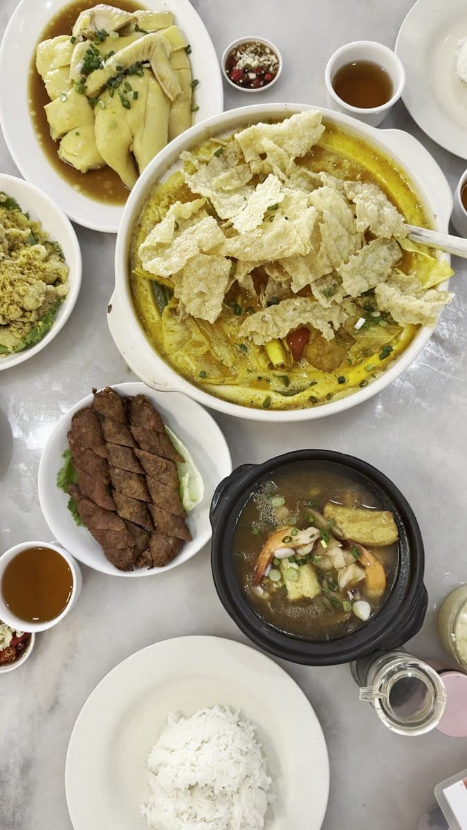 A table seen from above: sliced chicken, a claypot of curry topped with beancurd skin, fried meat rolls, a prawn and tofu soup, white rice and tea